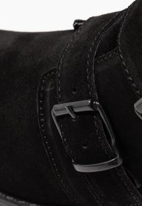 Botín negro de ante con una superficie texturizada, un acento de hebilla prominente y detalles cosidos a lo largo de los bordes. Hardware de alto contraste.