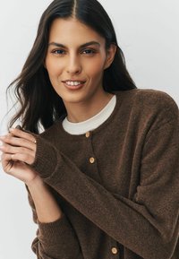 Hnědý pletený cardigan vyrobený z měkkého materiálu, s kulatým výstřihem, žebrovanými manžetami a světlými dřevěnými knoflíky. Modelovy ruce jsou umístěny na látce.