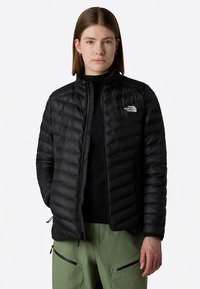 Czarna kurtka puchowa o falistym fakturze, z zamkiem błyskawicznym i wysokim kołnierzem. Na lewej piersi znajduje się logo, kurtka jest lekka.