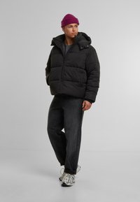 Svart pufferjakke med høy krage, glidelås og frontlommer, kombinert med mørke denimbukser og grå-hvite sneakers. En lilla lue er brukt.
