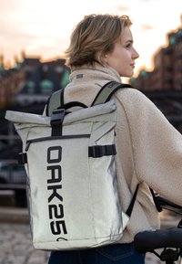 Mochila plateada de tipo roll-top con un gran logo negro "OAK25". Cuenta con correas ajustables, bolsillo frontal con cremallera y una tela resistente.