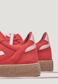 Baskets en suède rouge avec des accents roses, présentant une semelle en caoutchouc marron texturée et un rembourrage prononcé autour de la cheville. Marque dorée sur le côté.