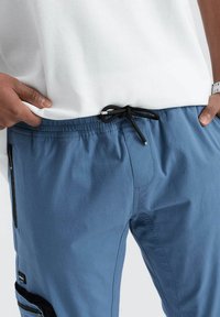 Pantalons bleu avec cordon de serrage, au tissu lisse, dotés de poches latérales à fermeture éclair et d'une taille élastique avec un lien noir.