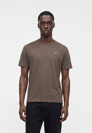 Ung mand iført en ensfarvet brun kortærmet t-shirt med et lille hvidt logo på brystet, stående foran en lys ensfarvet baggrund.