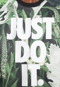 Grön sweatshirt med bladmönster och djärv vit text som säger "JUST DO IT." Har en rund svart krage och en strukturerad tyg.
