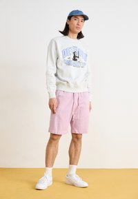 Light grey sweatshirt with blue graphic text, paired with pink shorts. White sneakers and a denim cap complete the outfit.