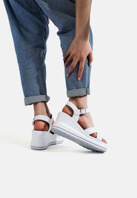 Sandali con zeppa bianchi con design a strisce e fibbia regolabile, indossati con jeans blu denim arrotolati. La suola presenta un motivo di grip testurizzato.