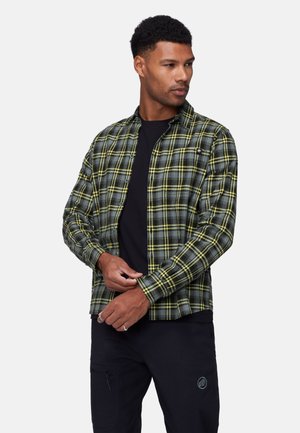 Mand justerer manchet på grøn og grå ternet skjorte, der bæres over sort t-shirt og sorte bukser mod en ensfarvet hvid baggrund.