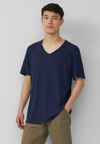 Granatowa koszulka typu v-neck z krótkim rękawem, wykonana z teksturowanej tkaniny. Nosi casualowo z rękami w kieszeniach, w połączeniu z khaki szortami.