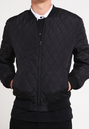 Homme portant une veste bomber matelassée noire par-dessus une chemise blanche à col, les mains dans les poches de la veste.