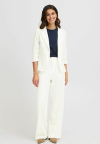 Blazer blanc et pantalon large assorti sur une chemise bleu marine, fabriqués en tissu lisse. Texture douce avec poches avant et design simple.