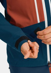 Blue and rust-colored jacket with a zip closure, featuring a velcro cuff adjustment and smooth fabric texture.