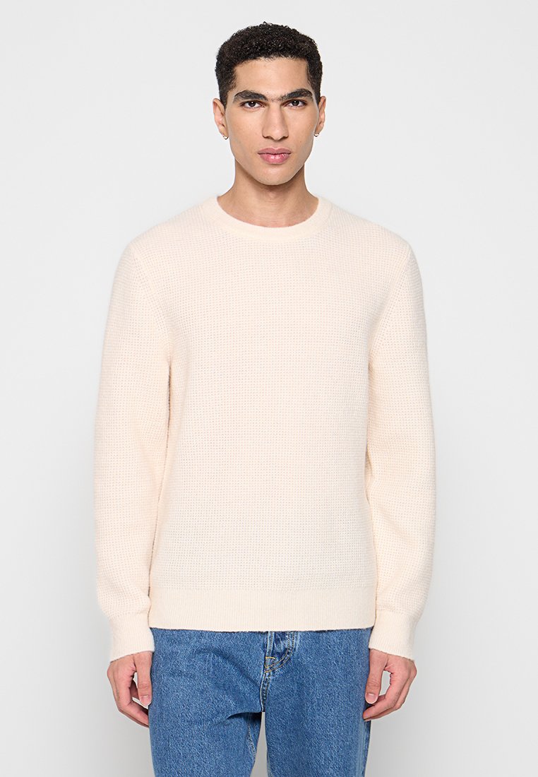 Hellbeiger strukturierter Pullover mit rundem Halsausschnitt und langen Ärmeln, kombiniert mit blauen Jeans. Einfaches Design, ohne Muster oder Akzente.