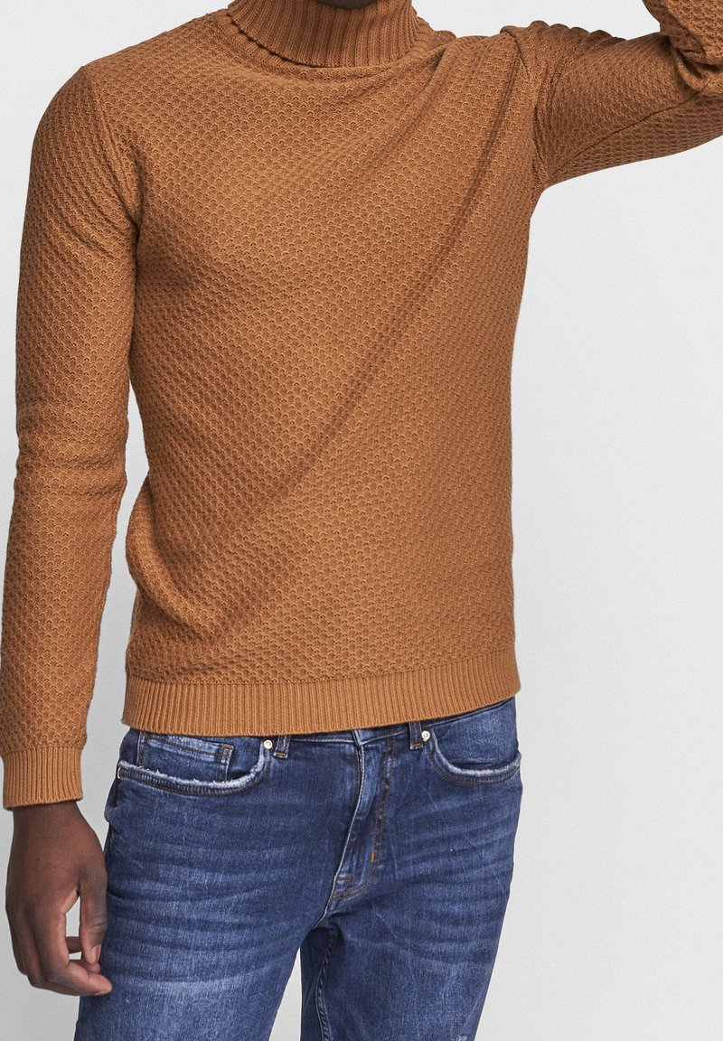 Pull marron en tricot avec un col montant, présentant un motif texturé. Porté avec un jean denim bleu, affichant une coupe décontractée.