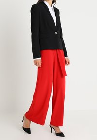 Kvinna i svart kavaj, vit skjorta, röda vida byxor och svarta spetsiga högklackade skor, stående mot en enkel bakgrund.