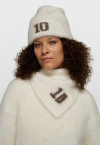 Mujer con cabello rizado lleva un gorro y bufanda de punto blancos a juego, ambos con el número "10" en hilo oscuro.