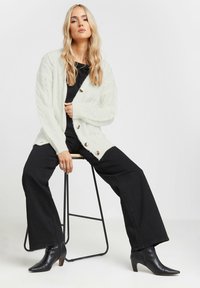 Cardigan bianco oversize a maglia grossa con grandi bottoni in legno, abbinato a pantaloni neri a gamba larga e stivali alla caviglia neri con tacco.