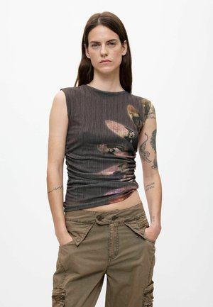 Vrouw met lang bruin haar, dragen een mouwloos donkergrijs top met bloemenprint en bruine cargobroek, handen in de zakken, neutrale uitdrukking.