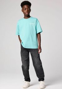 Ljus turkos bomullströja med oversize passform och vit logotryck, kombinerad med mörkgrå jeans och vita sneakers.