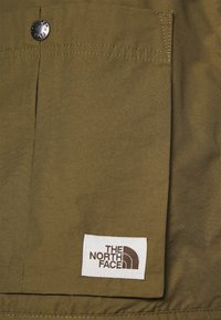 Grön tygficka med en silverfärgad tryckknapp och en vit rektangulär etikett som visar The North Face-logotypen i brunt.