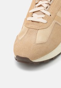 Beige mockaskons med texturerad ovandel, crèmefärgad mellansula och brun sula. Har rund snörning och rundad tå för en klassisk stil.