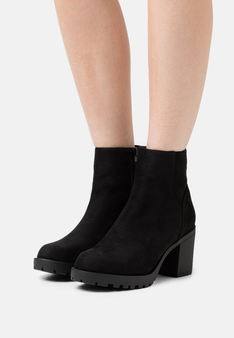 Jambes portant des bottines en daim noir à talons carrés et fermetures éclair latérales, posées sur une surface gris clair.