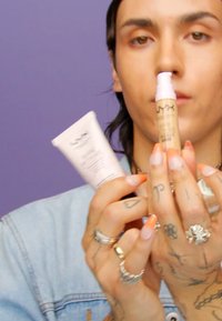 Två NYX kosmetikprodukter hålls: en klar, pumpflaska serum och en rosa, tubprimer. Bakgrunden är lila. Händer prydda med ringar.