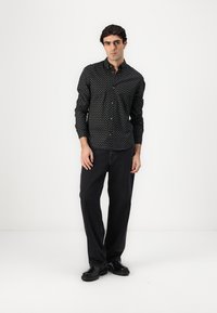 Schwarzes Hemd mit weißen Punkten, leichtes Material, taillierte Passform, kombiniert mit dunklen Jeans und schwarzen Schuhen.