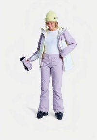 Veste de ski violette avec des accents blancs et jaunes, associée à un pantalon violet assorti. Le modèle tient des gants noirs et porte un bonnet jaune clair.