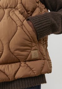Brown quilted vest with a texture of padded diamond shapes. Features a left-side pocket and a branded patch. Brown ribbed sweater underneath.