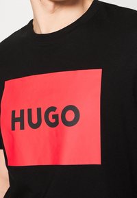 Schwarzes T-Shirt mit kurzen Ärmeln, das ein großes rotes rechteckiges Grafikdesign zeigt, in dem das Wort "HUGO" in fetten schwarzen Buchstaben zentriert ist.