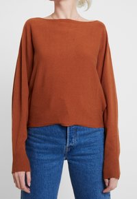 Übergroßer Pullover in verbranntem Orange aus weichem Stoff, mit weitem Halsausschnitt und gerippten Bündchen, kombiniert mit blauen High-Waist-Jeans.