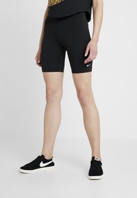Person som bär svarta Nike cykelshorts och svarta Nike sneakers, står med benen något isär på en vit bakgrund.
