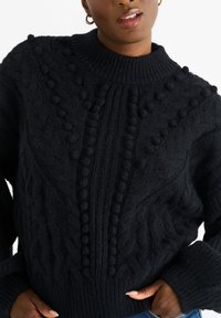 Maglione lavorato a maglia nera con colletto alto, caratterizzato da motivi a treccia e dettagli con pompon. Il materiale appare morbido e strutturato, con polsini a costine.