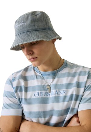 Giovane che indossa un cappello a secchiello grigio e una t-shirt a righe blu e bianche di Guess Jeans, con le braccia incrociate, guarda verso il basso con una collana d'argento.