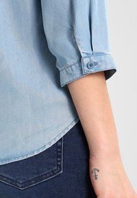 Hellblaues Denimhemd mit hochgekrempelten Ärmeln, das mit einem Knopfdetails am Bündchen versehen ist. Tragen mit dunkelblauen Jeans, die ein Tattoo am Handgelenk zeigen.