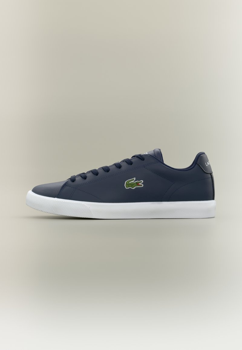 Sapatilha de cano baixo azul-marinho com sola branca, fecho de atacadores à frente, e logótipo de crocodilo verde de lado.