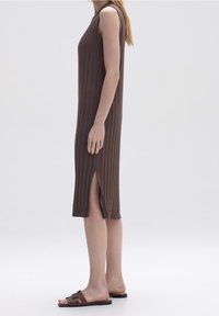 Mujer de pie con un vestido midi de canalé marrón sin mangas y una abertura lateral, usando sandalias planas marrones sobre un fondo claro.
