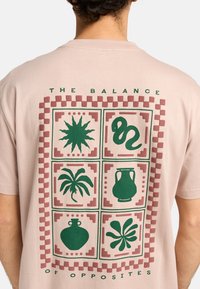 Lichtroze katoenen t-shirt met een groene grafische print op de achterkant. Het ontwerp omvat vier kwadranten met verschillende vormen en patronen.