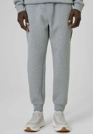 Osoba ubrana w jasnoszare spodnie dresowe i białe trampki z gumowymi podeszwami, stojąca na jednolitym jasnym tle.