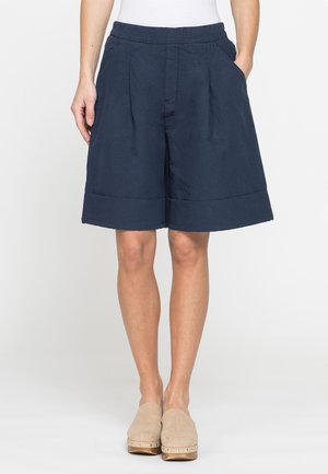 Donna che indossa pantaloncini blu navy a gamba larga con tasche laterali e zoccoli beige slip-on con suole in legno e borchie.