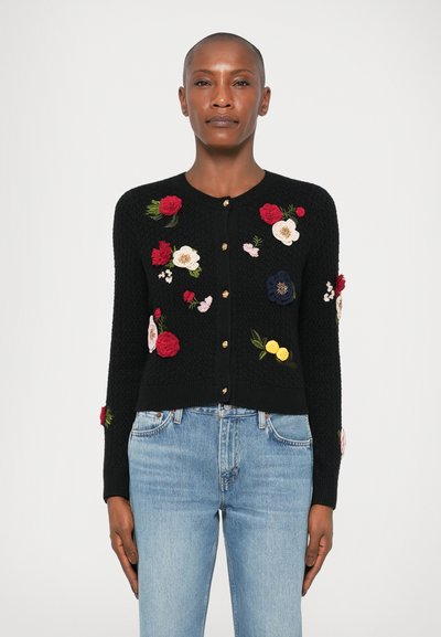 Cardigan en tricot noir avec broderies florales colorées, boutons dorés et poignets côtelés. Porté avec un jean en denim bleu clair.
