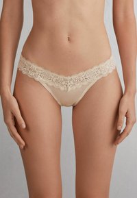 Beige kanten string met bloemenpatroon en golvende randen, voorzien van doorschijnende stof en een lage taille, die minimale bedekking biedt.