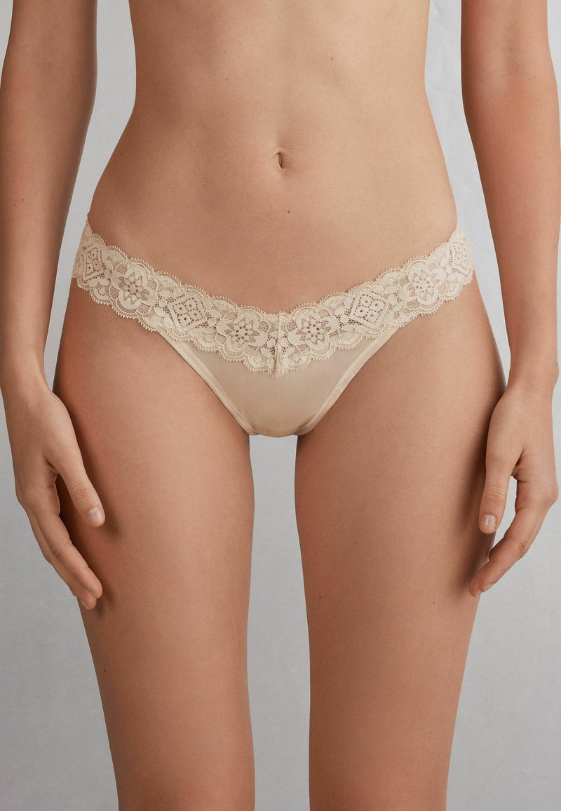 Beige kanten string met bloemenpatroon en golvende randen, voorzien van doorschijnende stof en een lage taille, die minimale bedekking biedt.