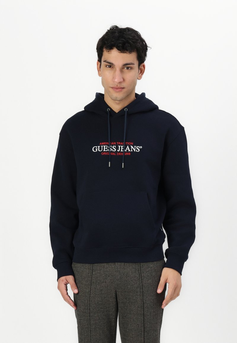 Temno modra kapuca s sprednjim žepom, kapuco z vrvico in izvezenim logotipom "GUESS JEANS" v rdeči in beli barvi čez prsi.