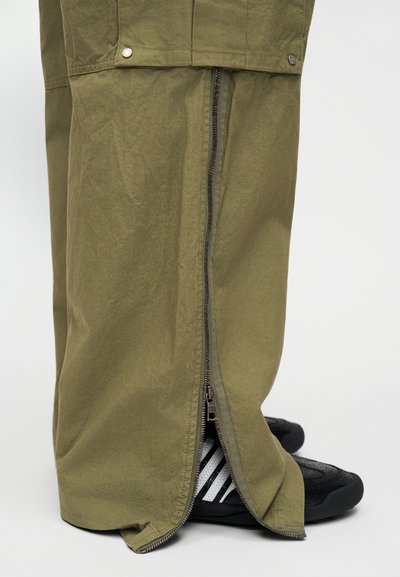 Χαλαρά παντελόνια σε ελιά πράσινο με μακρύ φερμουάρ στο πλάι και ασημένια κουμπιά τύπου snap, φορεμένα πάνω από μαύρα παπούτσια με λευκές ρίγες.
