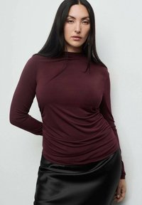 Burgundijska majica z dolgimi rokavi in visokim ovratnikom, z nabranim teksturo, v kombinaciji z elegantno črno satensko krilo, ki poudarja gladke materiale in fitted oblike.