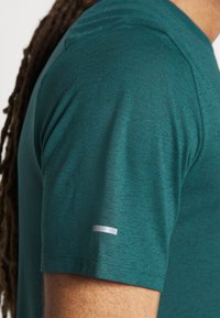 Teal short-sleeve shirt with a textured finish, featuring a small white accent on the sleeve. Fabric appears lightweight and breathable.