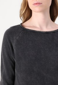 Tamsiai pilkas džemperis su plačiu apykaklės iškirpimu, raglanų stiliaus rankovėmis ir minkštu paviršiumi; turi šiek tiek suplėšytą apykaklės kraštą.