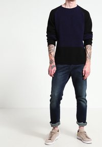 Man som bär en marinblå och svart tröja, uppvikta mörkblå jeans, beige sneakers och synliga tatueringar på båda underarmarna och händerna.